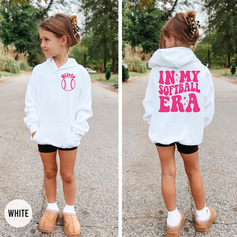 May include: White hoodie with a front pocket and a hood. The front features the name "Olivia" above a pink softball graphic. The back of the hoodie displays the text "IN MY SOFTBALL ERA" in pink. The hoodie is worn by a person.