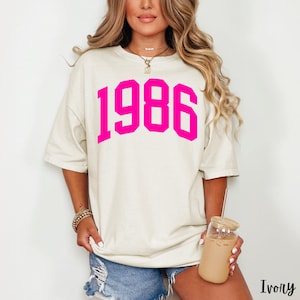 May include: Ivory-colored t-shirt with the year "1986" printed in bright pink. The shirt has a relaxed fit and short sleeves. The model is holding a glass of iced coffee.