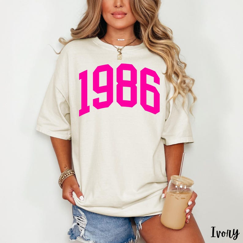 May include: Ivory-colored t-shirt with the year "1986" printed in bright pink. The shirt has a relaxed fit and short sleeves. The model is holding a glass of iced coffee.