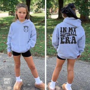 May include: Light grey hoodie with a front pocket and a baseball graphic. The front of the hoodie has the name "Olivia" above the baseball graphic. The back of the hoodie says "IN MY SOFTBALL ERA" in black lettering.