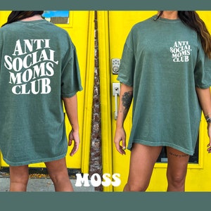 May include: A green oversized t-shirt with the text "ANTI SOCIAL MOMS CLUB" in white. The shirt is being worn by a person standing in front of a yellow door.