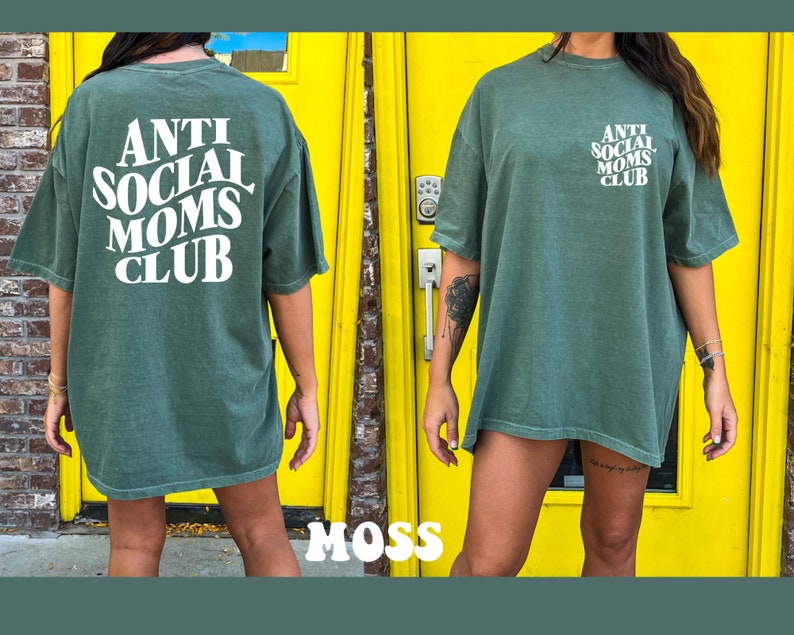 May include: A green oversized t-shirt with the text "ANTI SOCIAL MOMS CLUB" in white. The shirt is being worn by a person standing in front of a yellow door.