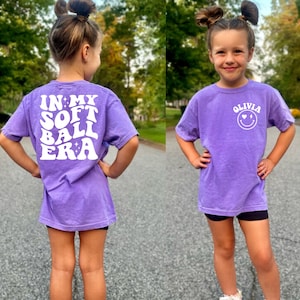 May include: Purple short-sleeved t-shirt with white text on the back that says "In My Softball Era". The front of the shirt has white text that says "Olivia" with a smiley face with hearts for eyes and a lightning bolt for a mouth.
