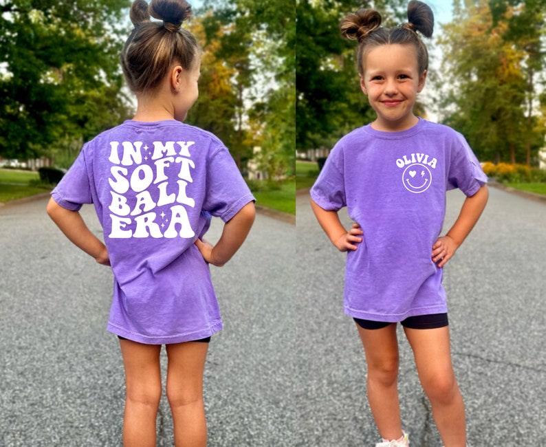 May include: Purple short-sleeved t-shirt with white text on the back that says "In My Softball Era". The front of the shirt has white text that says "Olivia" with a smiley face with hearts for eyes and a lightning bolt for a mouth.