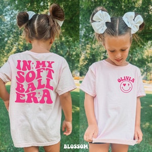 May include: Pink t-shirt with a pink graphic that says "In My Softball Era" on the back and a pink graphic with a smiley face and the text "Olivia" on the front.