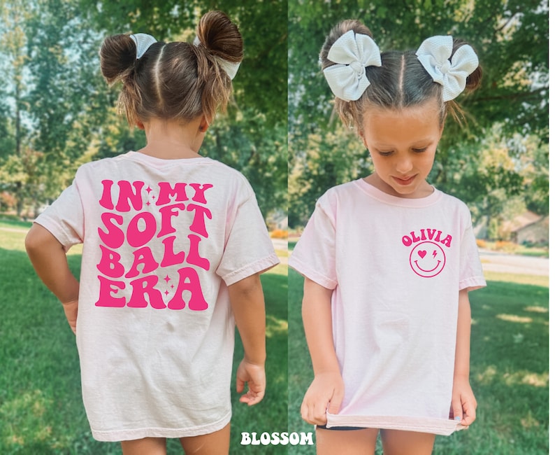 May include: Pink t-shirt with a pink graphic that says "In My Softball Era" on the back and a pink graphic with a smiley face and the text "Olivia" on the front.