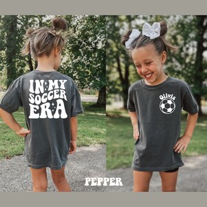 May include: A dark gray t-shirt with white text that says "In My Soccer Era" on the back and a white soccer ball with the name "Olivia" on the front.