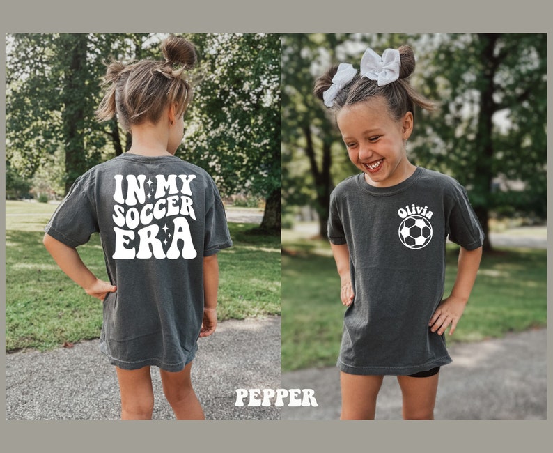 May include: A dark gray t-shirt with white text that says "In My Soccer Era" on the back and a white soccer ball with the name "Olivia" on the front.