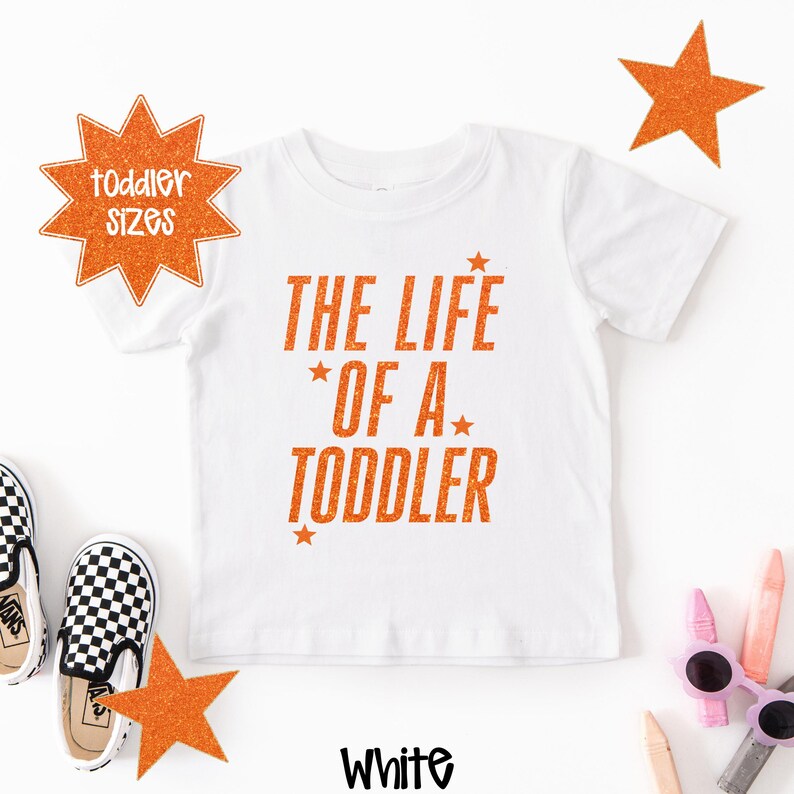 Pu&ograve; includere: T-shirt bianco per bambini piccoli con la scritta "THE LIFE OF A TODDLER" in glitter arancione. La maglietta ha girocollo e maniche corte. Sono presenti anche stelle arancioni e una grafica a stella con "toddler sizes".