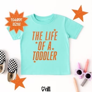 Pu&ograve; includere: Una maglietta turchese per bambini piccoli con la scritta arancione "THE LIFE OF A TODDLER". Il design include stelle arancioni e un paio di scarpe a scacchi bianchi e neri. La parola "Chill" &egrave; in basso nell'immagine.