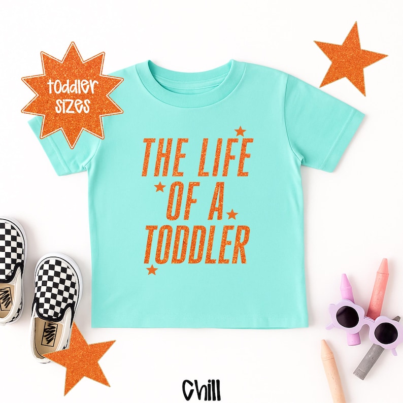 Pu&ograve; includere: Una maglietta turchese per bambini piccoli con la scritta arancione "THE LIFE OF A TODDLER". Il design include stelle arancioni e un paio di scarpe a scacchi bianchi e neri. La parola "Chill" &egrave; in basso nell'immagine.