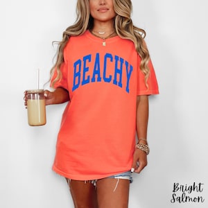 May include: Bright salmon-colored t-shirt with the word "BEACHY" in bold blue letters. The shirt is a short-sleeved, casual style. The words "Bright Salmon" are written in a script font in the bottom right corner.