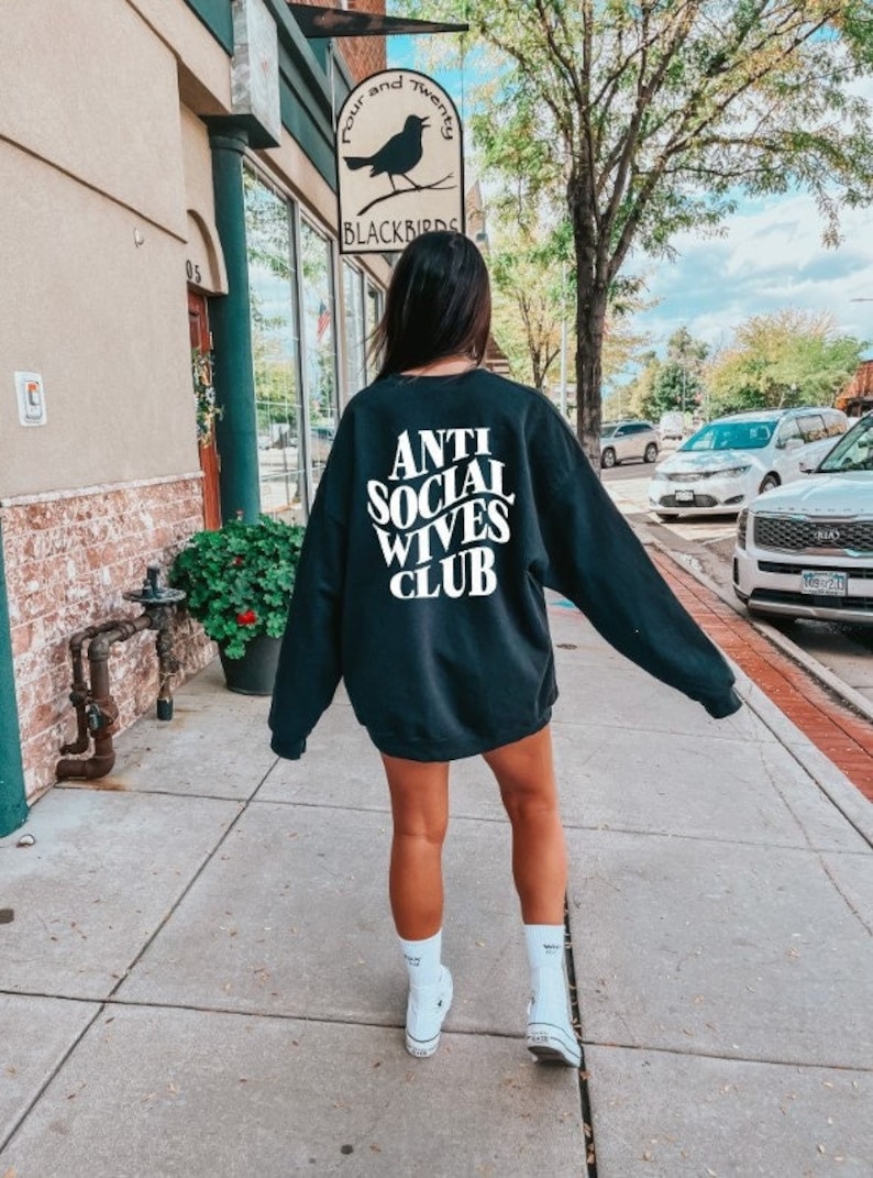 Puede incluir: Una mujer lleva una sudadera negra con el texto "Anti Social Wives Club" en letras blancas. Lleva calcetines blancos y zapatillas blancas. Est&aacute; caminando por una acera frente a un edificio.