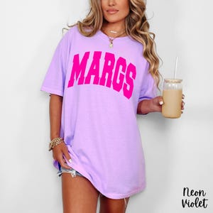 May include: A light purple t-shirt with the word "MARGS" in bright pink block letters across the front. The shirt is a relaxed fit and has short sleeves. The text "Neon Violet" is in the bottom right corner.