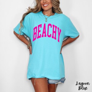 May include: A light blue t-shirt with the word "BEACHY" in bright pink block letters across the chest. The shirt is a relaxed fit and has short sleeves. The text "Lagoon Blue" is written in cursive at the bottom right.