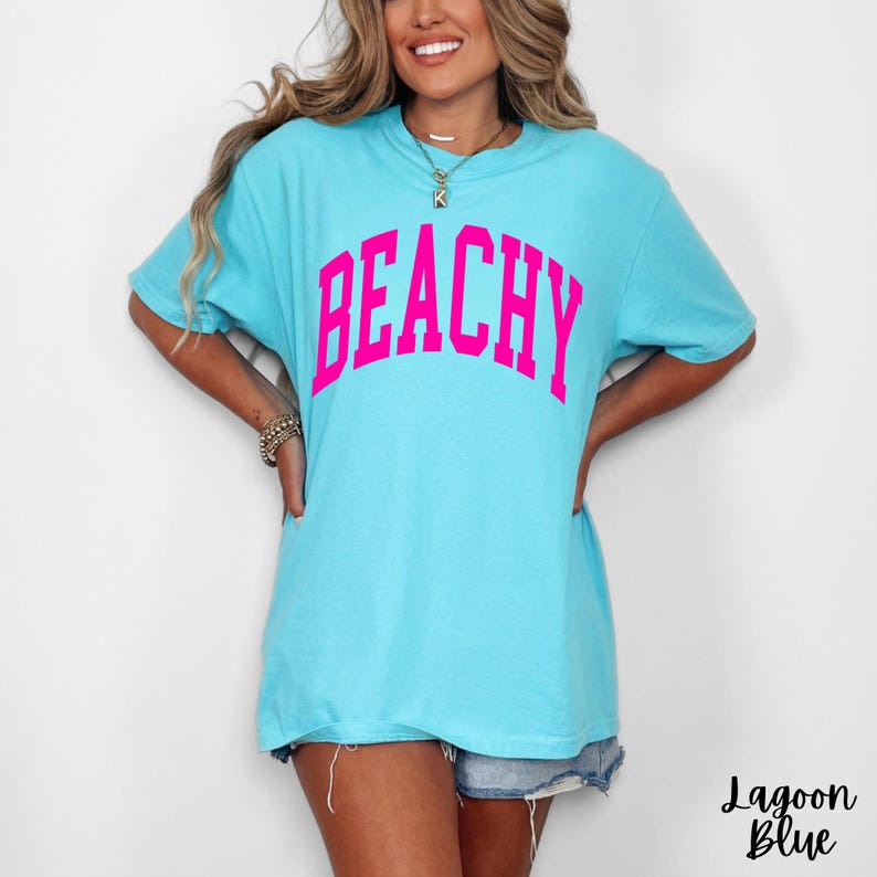 May include: A light blue t-shirt with the word "BEACHY" in bright pink block letters across the chest. The shirt is a relaxed fit and has short sleeves. The text "Lagoon Blue" is written in cursive at the bottom right.