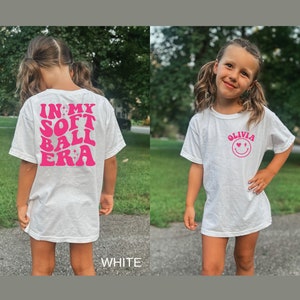 May include: White t-shirt with pink text that says "In My Soft Ball Era" on the back and a pink smiley face with the text "Olivia" on the front.