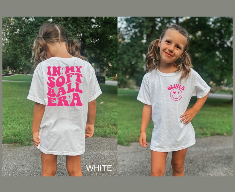May include: White t-shirt with pink text that says "In My Soft Ball Era" on the back and a pink smiley face with the text "Olivia" on the front.