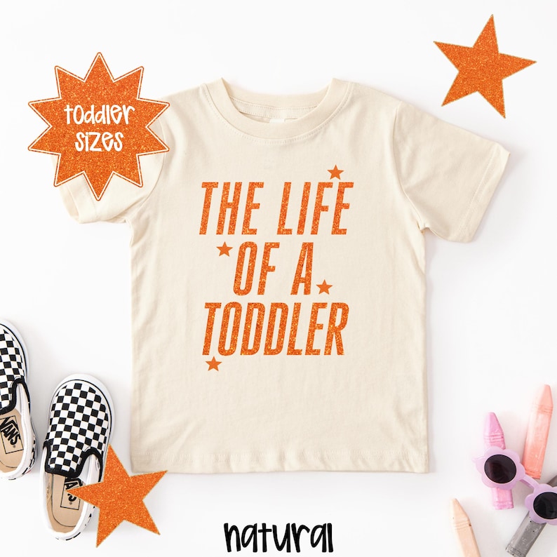 Pu&ograve; includere: Una maglietta per bambini piccoli di colore naturale con la scritta "THE LIFE OF A TODDLER" in arancione. Il design include stelle arancioni. Le parole "toddler sizes" sono a forma di stella arancione. La parola "natural" &egrave; in basso.