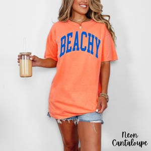 May include: Neon cantaloupe-colored t-shirt with the word "BEACHY" in blue block letters. The shirt is paired with denim shorts. A glass tumbler with a straw is held in the left hand.