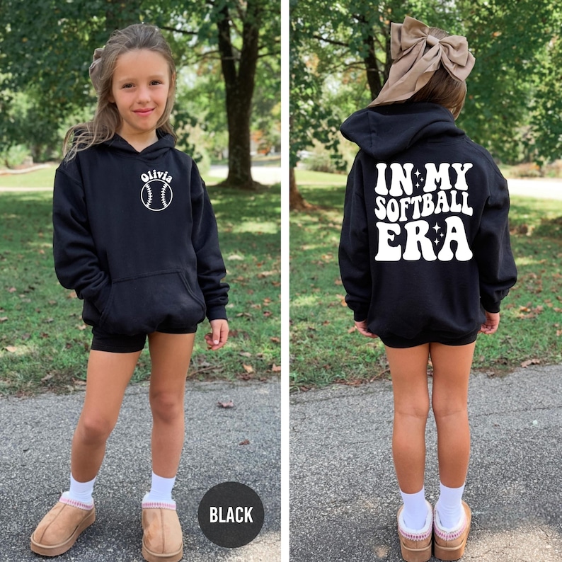 May include: Black hoodie with a front pocket and a white softball graphic with the name "Olivia". The back of the hoodie says "IN MY SOFTBALL ERA" in white lettering. The hoodie is worn with black shorts and white socks.