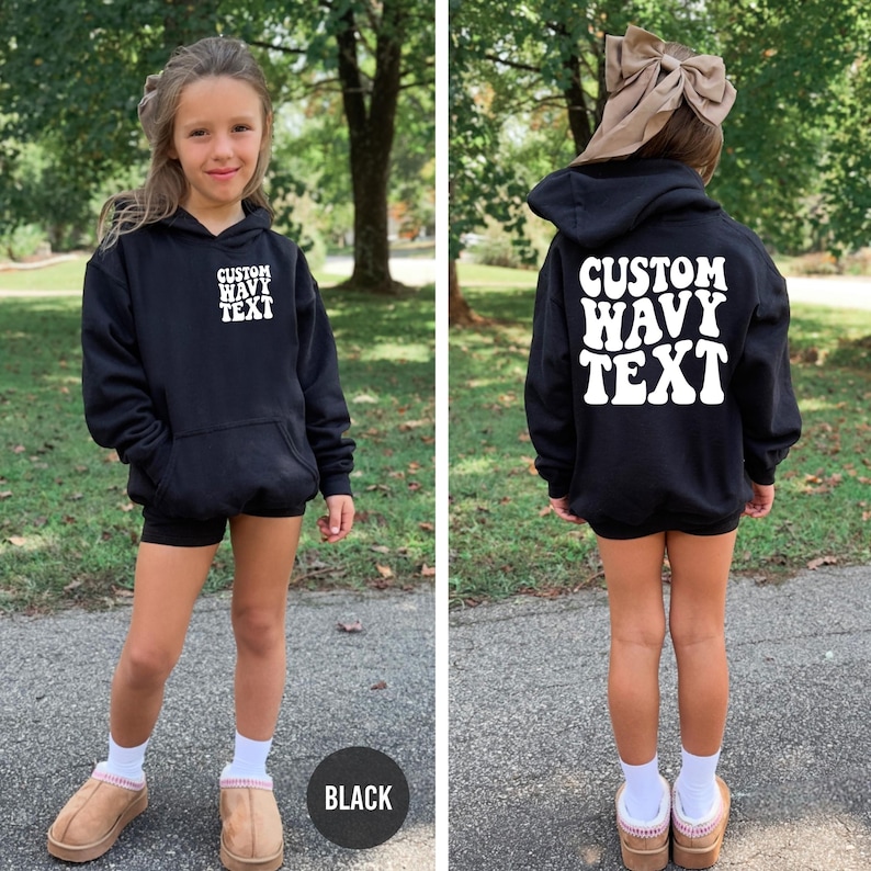 Puede incluir: Sudadera con capucha negra con el texto "CUSTOM WAVY TEXT" en blanco en la parte delantera y trasera. La sudadera tiene capucha y bolsillo delantero. La modelo lleva shorts negros, calcetines blancos y zapatillas marrones.