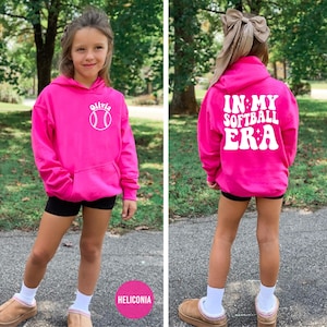 May include: A bright pink hoodie with a front pocket and white text. The front of the hoodie has the name "Olivia" and a softball graphic. The back of the hoodie says "IN MY SOFTBALL ERA" in white text. The hoodie is worn by a person.