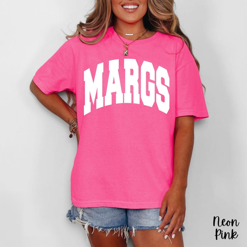May include: Neon pink t-shirt with the word "MARGS" in large white block letters across the chest. The shirt is a vibrant, solid color and has short sleeves. The text "Neon Pink" is in the bottom right corner.