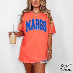 May include: Bright salmon-colored t-shirt with the word "MARGS" in blue block letters. The shirt is a crew neck style and appears to be made of a soft material. The model is wearing denim shorts and holding a glass with a straw.