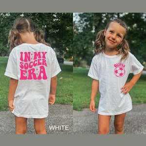 May include: White t-shirt with pink text that says "In My Soccer Era" on the back and a pink soccer ball with the name "Olivia" on the front.