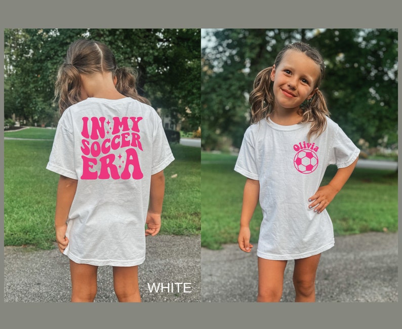 May include: White t-shirt with pink text that says "In My Soccer Era" on the back and a pink soccer ball with the name "Olivia" on the front.