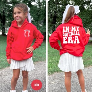 May include: A red hooded sweatshirt with the text "Olivia" and a baseball graphic. The back of the sweatshirt says "IN MY SOFTBALL ERA" in white lettering. The person is wearing a white pleated skirt.