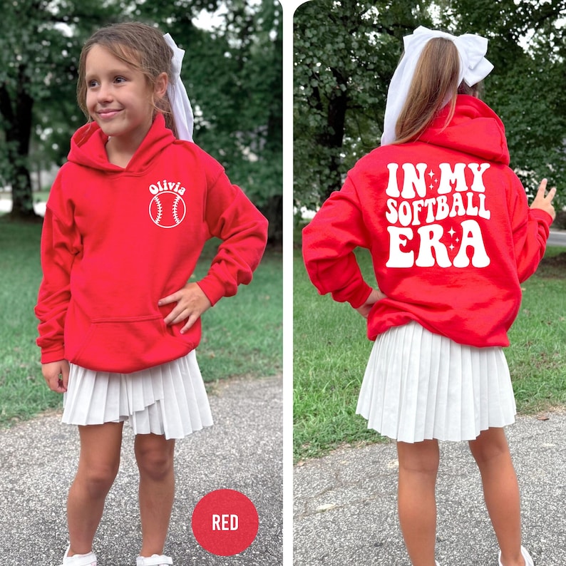 May include: A red hooded sweatshirt with the text "Olivia" and a baseball graphic. The back of the sweatshirt says "IN MY SOFTBALL ERA" in white lettering. The person is wearing a white pleated skirt.