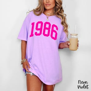 May include: A light purple t-shirt with the number "1986" printed in bright pink. The shirt is a relaxed fit and has short sleeves. The text "Neon Violet" is in the bottom right corner.