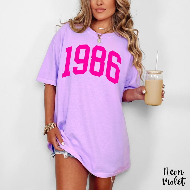 May include: A light purple t-shirt with the number "1986" printed in bright pink. The shirt is a relaxed fit and has short sleeves. The text "Neon Violet" is in the bottom right corner.
