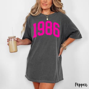 May include: Dark gray t-shirt with the year "1986" in bright pink block letters. The shirt has a relaxed fit and short sleeves. The word "Pepper" is printed in the bottom right corner.