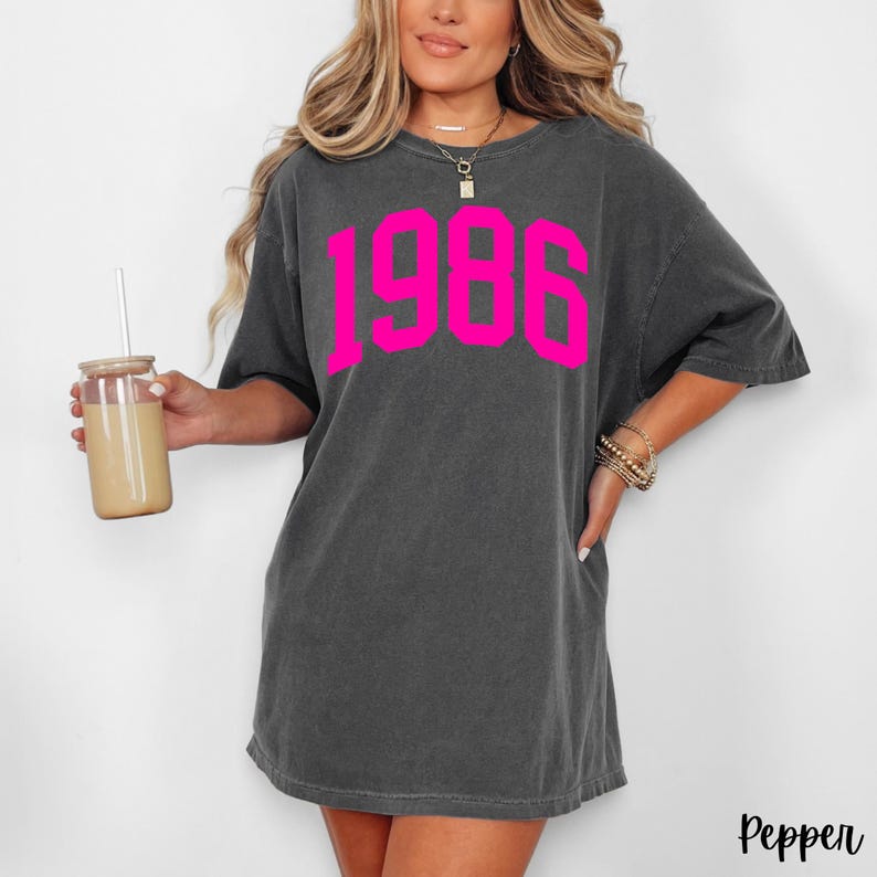 May include: Dark gray t-shirt with the year "1986" in bright pink block letters. The shirt has a relaxed fit and short sleeves. The word "Pepper" is printed in the bottom right corner.