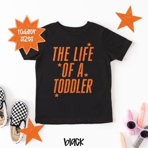Pu&ograve; includere: T-shirt nera per bambini piccoli con testo arancione "THE LIFE OF A TODDLER" e dettagli a stella. Una stella arancione mostra "toddler sizes". La parola "black" &egrave; scritta in un carattere corsivo.