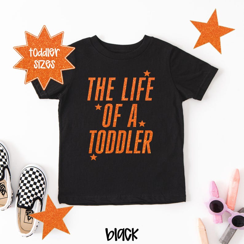 Pu&ograve; includere: T-shirt nera per bambini piccoli con testo arancione "THE LIFE OF A TODDLER" e dettagli a stella. Una stella arancione mostra "toddler sizes". La parola "black" &egrave; scritta in un carattere corsivo.