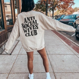 Puede incluir: Una sudadera beige con el texto "Anti Social Wives Club" impreso en la espalda en negro. La sudadera la lleva puesta una persona que est&aacute; de pie en una acera.