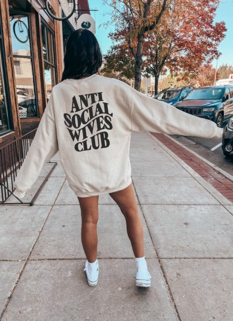 Puede incluir: Una sudadera beige con el texto "Anti Social Wives Club" impreso en la espalda en negro. La sudadera la lleva puesta una persona que est&aacute; de pie en una acera.