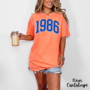 May include: A neon cantaloupe-colored t-shirt with the number "1986" in blue block letters. The shirt is paired with denim shorts. A glass tumbler with a straw is held in the left hand. The text "Neon Cantaloupe" is in the bottom right corner.