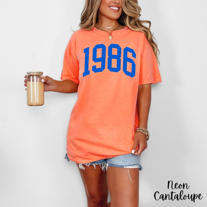 May include: A neon cantaloupe-colored t-shirt with the number "1986" in blue block letters. The shirt is paired with denim shorts. A glass tumbler with a straw is held in the left hand. The text "Neon Cantaloupe" is in the bottom right corner.