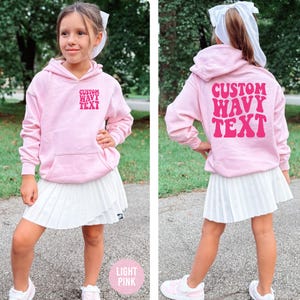 Puede incluir: Una sudadera con capucha rosa claro con el texto "CUSTOM WAVY TEXT" en la parte delantera y trasera. La sudadera tiene un bolsillo delantero y una capucha. La modelo lleva una falda plisada blanca y zapatillas rosas. El texto "LIGHT PINK" est&aacute; en la parte inferior.