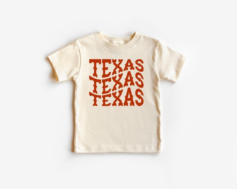 K&ouml;nnte beinhalten: Ein cremefarbenes T-Shirt mit kurzen &Auml;rmeln, auf dem das Wort "TEXAS" dreimal in einer roten, stilisierten Schrift wiederholt wird.