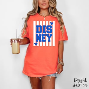 Może przedstawiać: Jasnor&oacute;żowa koszulka z napisem "DISNEY" w niebieskich literach, na białym tle w paski. Koszulka posiada małe sylwetki Myszki Miki. Odzież codzienna.