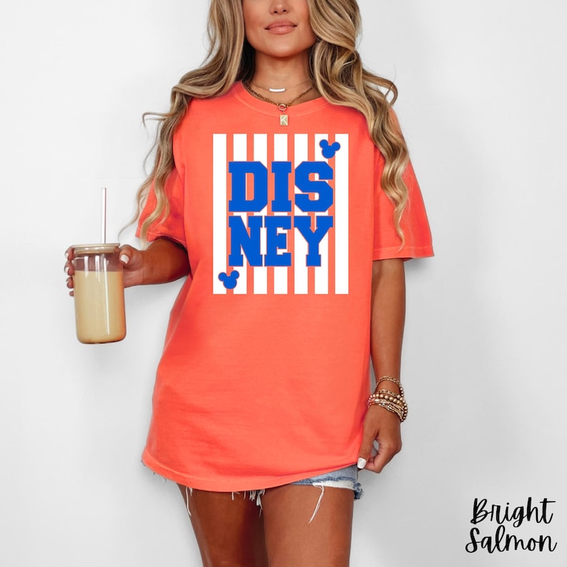 Może przedstawiać: Jasnor&oacute;żowa koszulka z napisem "DISNEY" w niebieskich literach, na białym tle w paski. Koszulka posiada małe sylwetki Myszki Miki. Odzież codzienna.