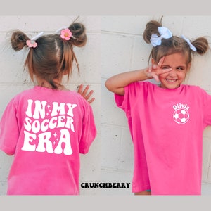 May include: A pink t-shirt with white text that says "In My Soccer Era" with stars. The back of the shirt has a white soccer ball with the name "Olivia" above it.