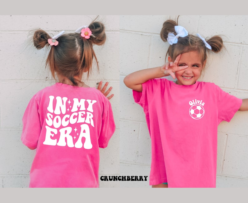 May include: A pink t-shirt with white text that says "In My Soccer Era" with stars. The back of the shirt has a white soccer ball with the name "Olivia" above it.
