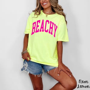 May include: A neon yellow t-shirt with the word "BEACHY" in bright pink block letters. The shirt is paired with denim shorts. The words "Neon Lemon" are in the bottom right corner.